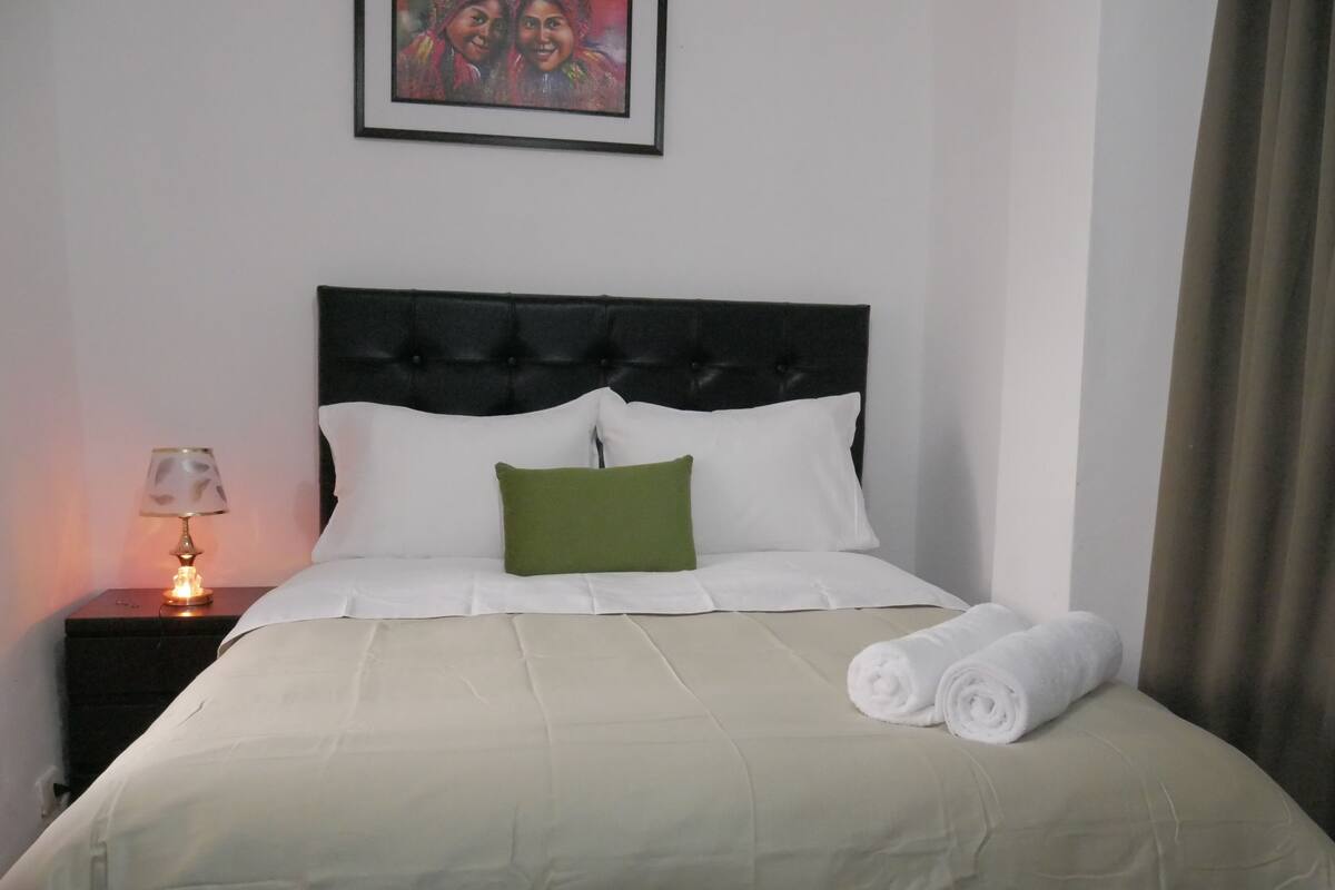 A neatly made bed with white bedding and a green pillow, a bedside lamp, and a framed picture on the wall.