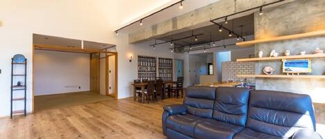 A spacious living area with a wooden floor, a leather sofa, a dining table with chairs, and a kitchen area with a brick backsplash.
