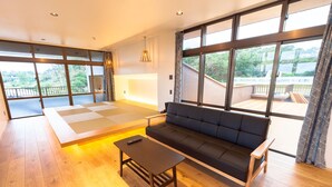 Amplia habitación con piso de madera, un sofá negro, una mesa de centro de madera y una zona con esteras de tatami. Amplias ventanas ofrecen vistas al exterior.