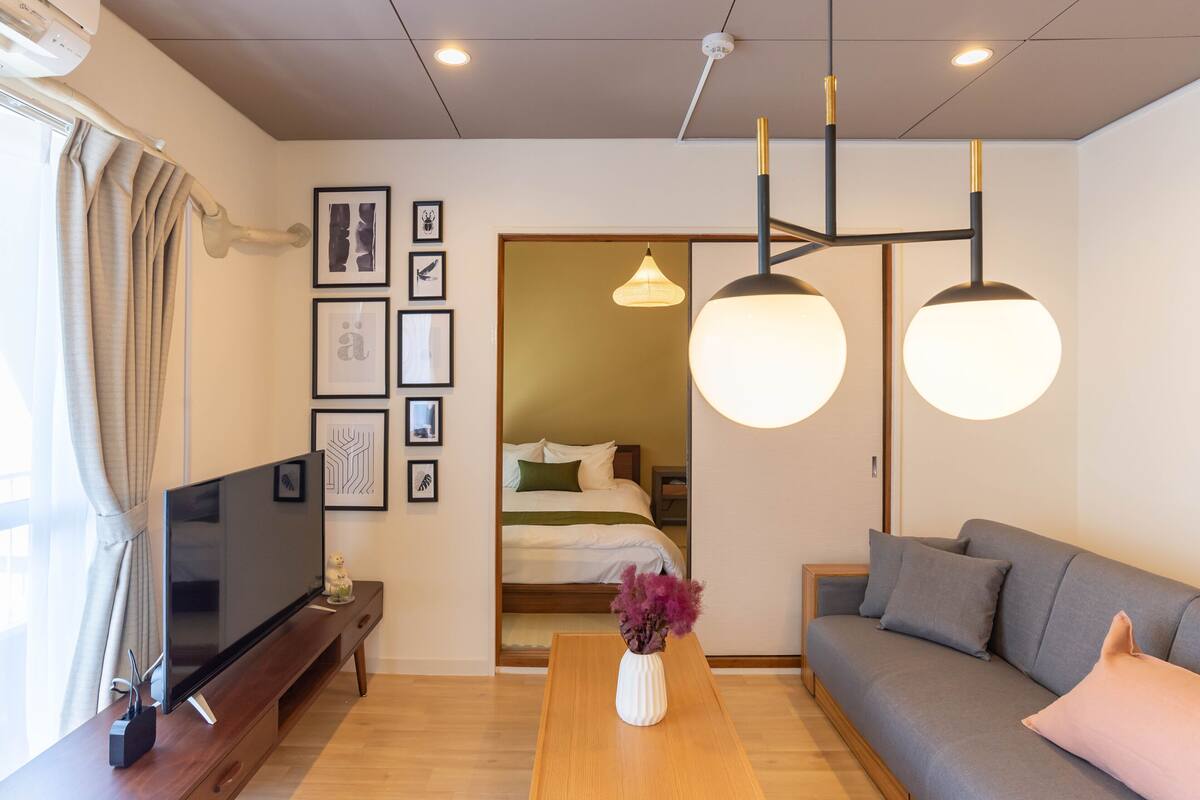 A modern living room with a flat-screen TV, wooden coffee table, grey sofa, and a large window with curtains.