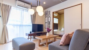 Ein modernes Wohnzimmer mit einer Couch, einem Hocker und einem Fernseher. Ein Holztischchen und ein Schreibtisch mit einem Computer stehen im Raum. Große Fenster mit Vorhängen sorgen für viel Licht.
