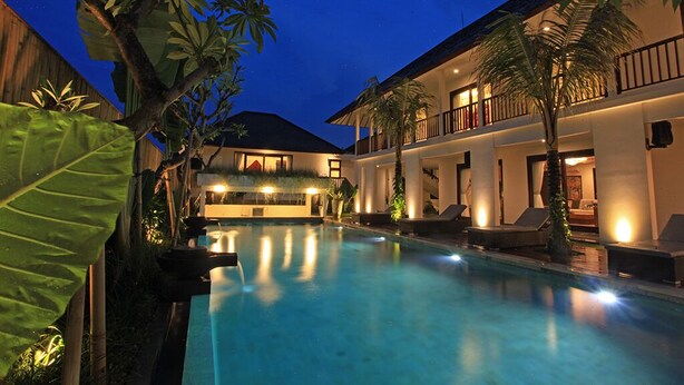 A pool area with lounge chairs, a building with balconies, and tropical plants. - Elite Cassia (Tibubeneng)