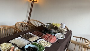 A dining table with various food items and a hot pot, surrounded by wooden chairs in a room with a wooden ceiling and a hanging light fixture.