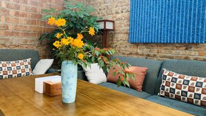 A cozy room with a brick wall, a blue patterned wall hanging, a wooden table with a vase of yellow flowers, and a grey sofa with patterned cushions.