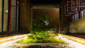 A nighttime view of a building with a brick wall, a red roof, and a small garden area with plants and a circular light fixture.