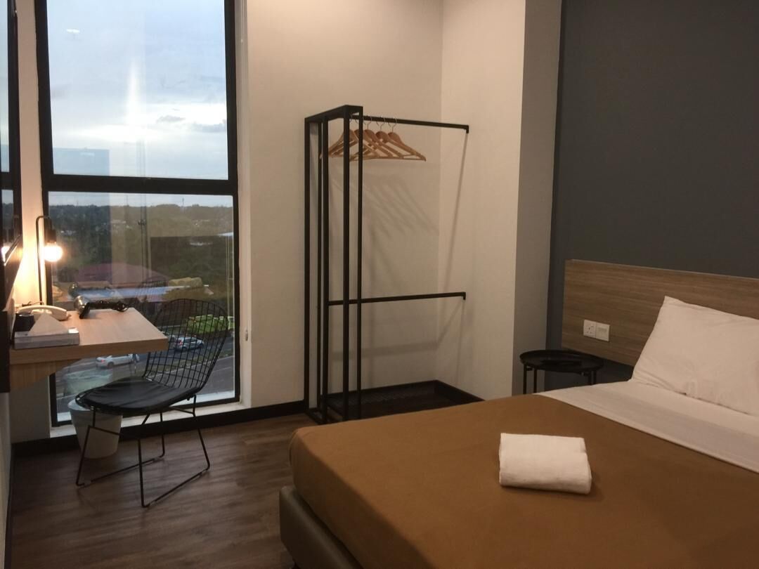 A hotel room with a bed, a chair, a desk with a lamp, a window, and a hanging clothes rack.