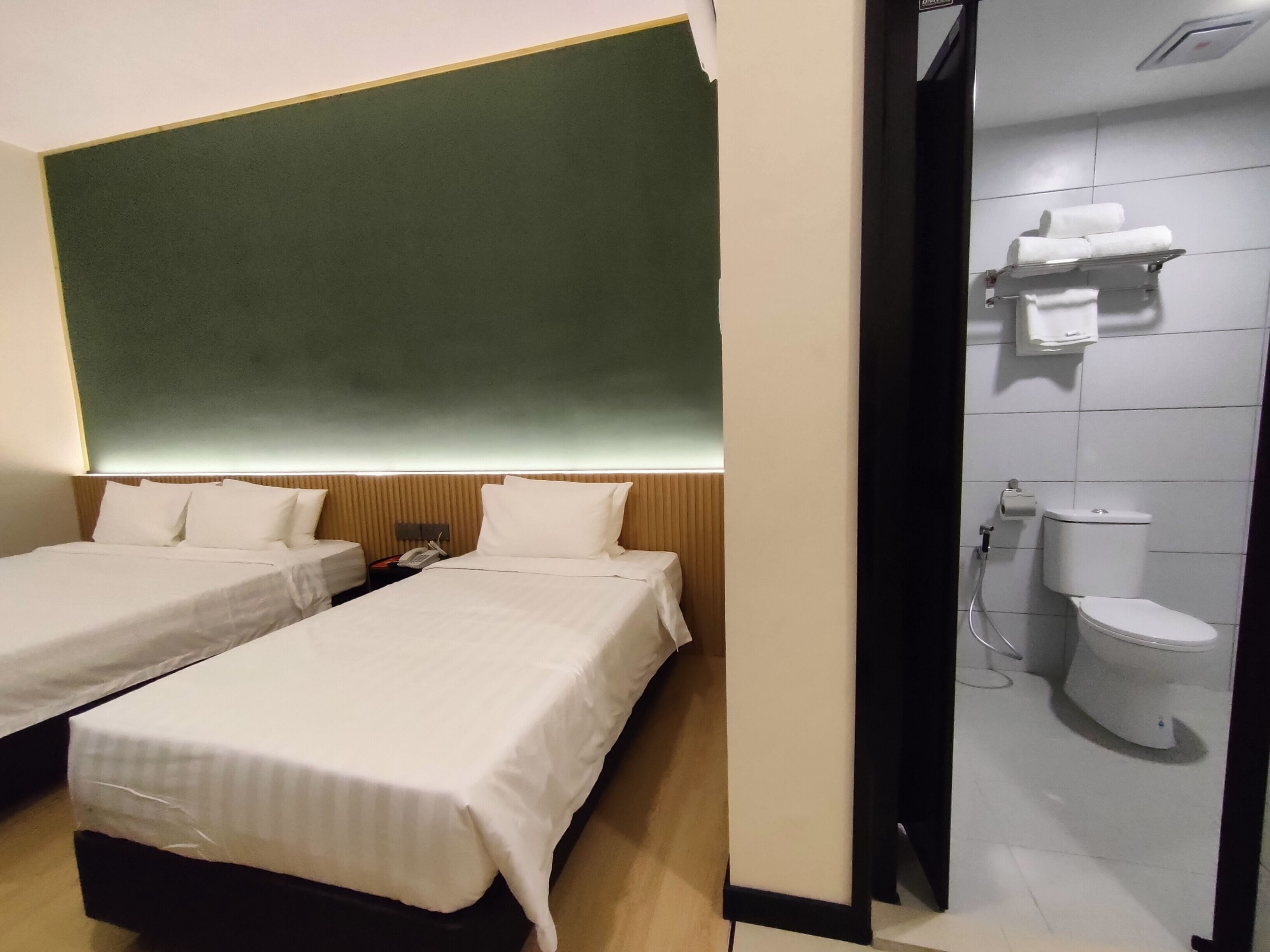 A hotel room with two beds, a green accent wall, and a bathroom with a toilet and towel rack.