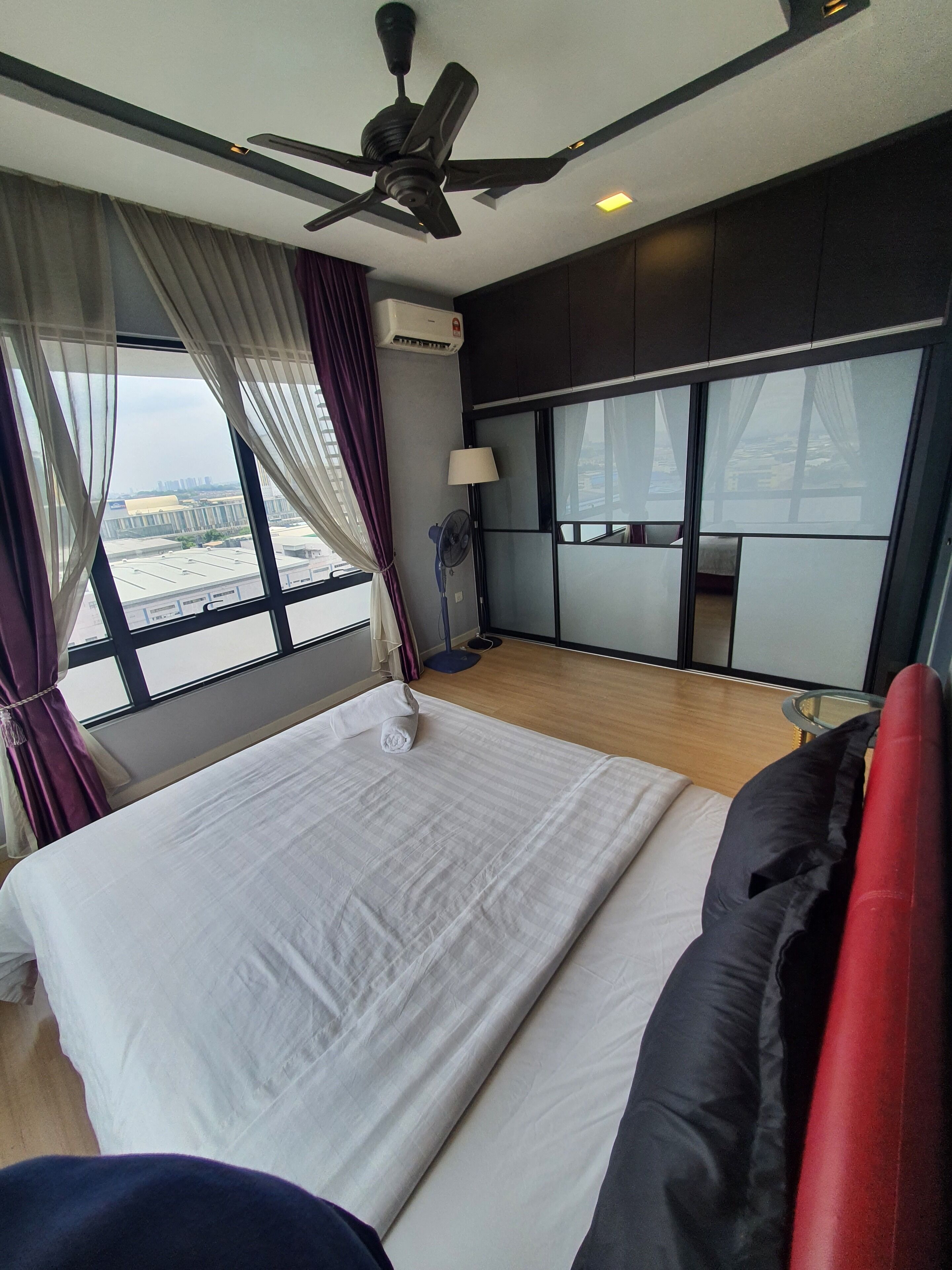 A hotel room with a bed, a ceiling fan, an air conditioning unit, a window with curtains, a bedside lamp, and a view of the cityscape.
