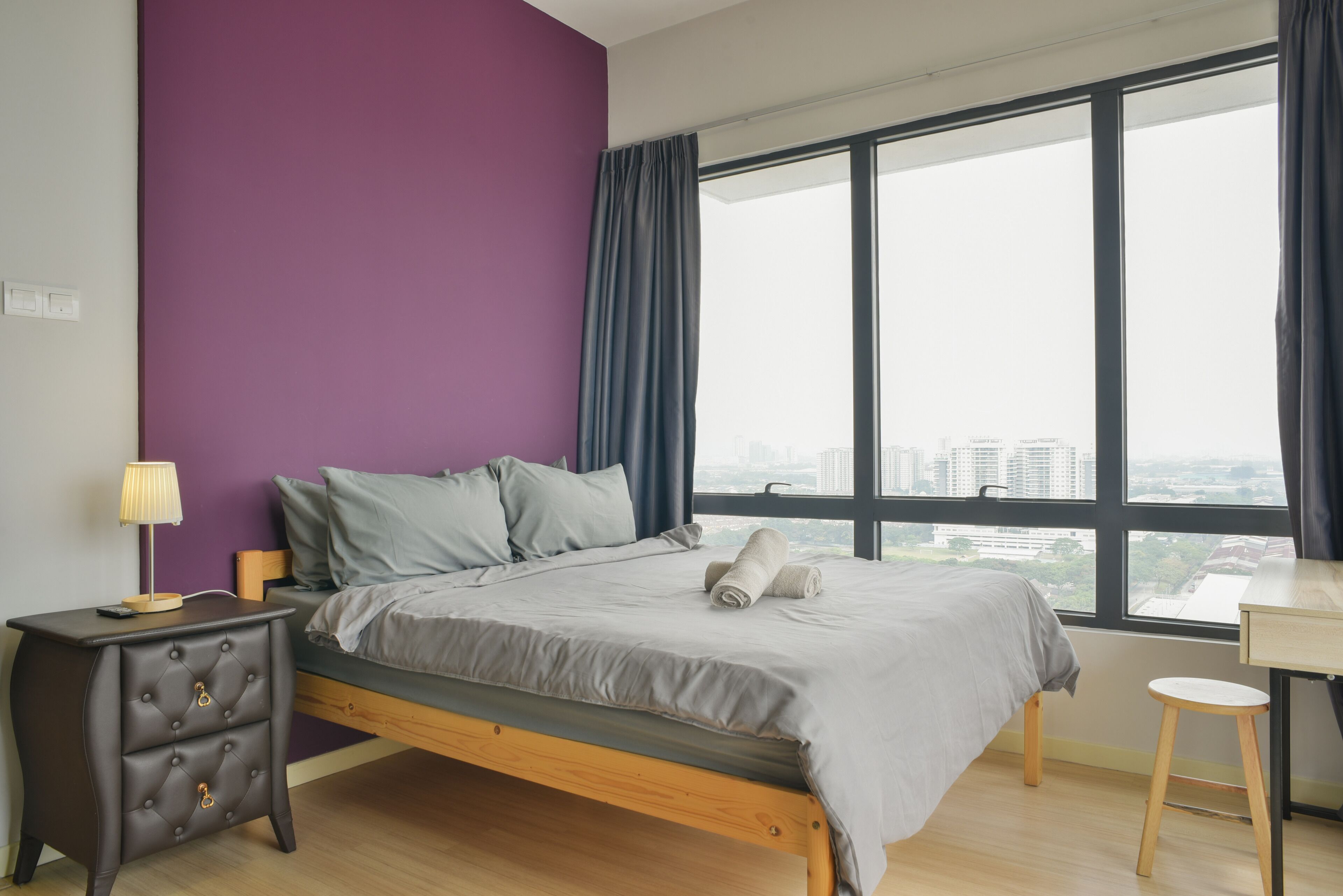 A bedroom with a bed, a nightstand, a lamp, a window with a view of buildings, and a small table.