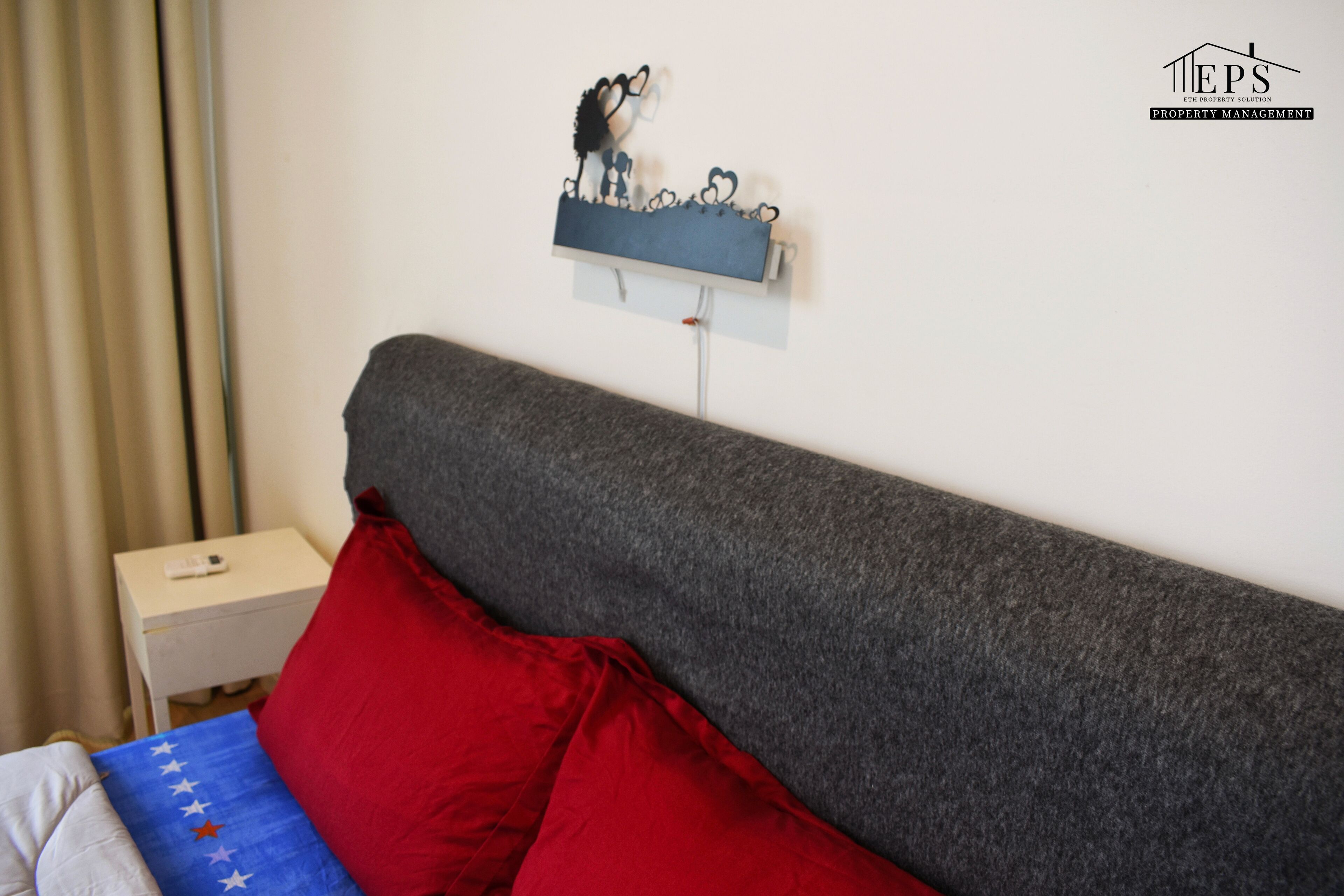 A bedroom with a grey headboard, red and blue pillows, a small bedside table, and a wall-mounted shelf with a decorative item.