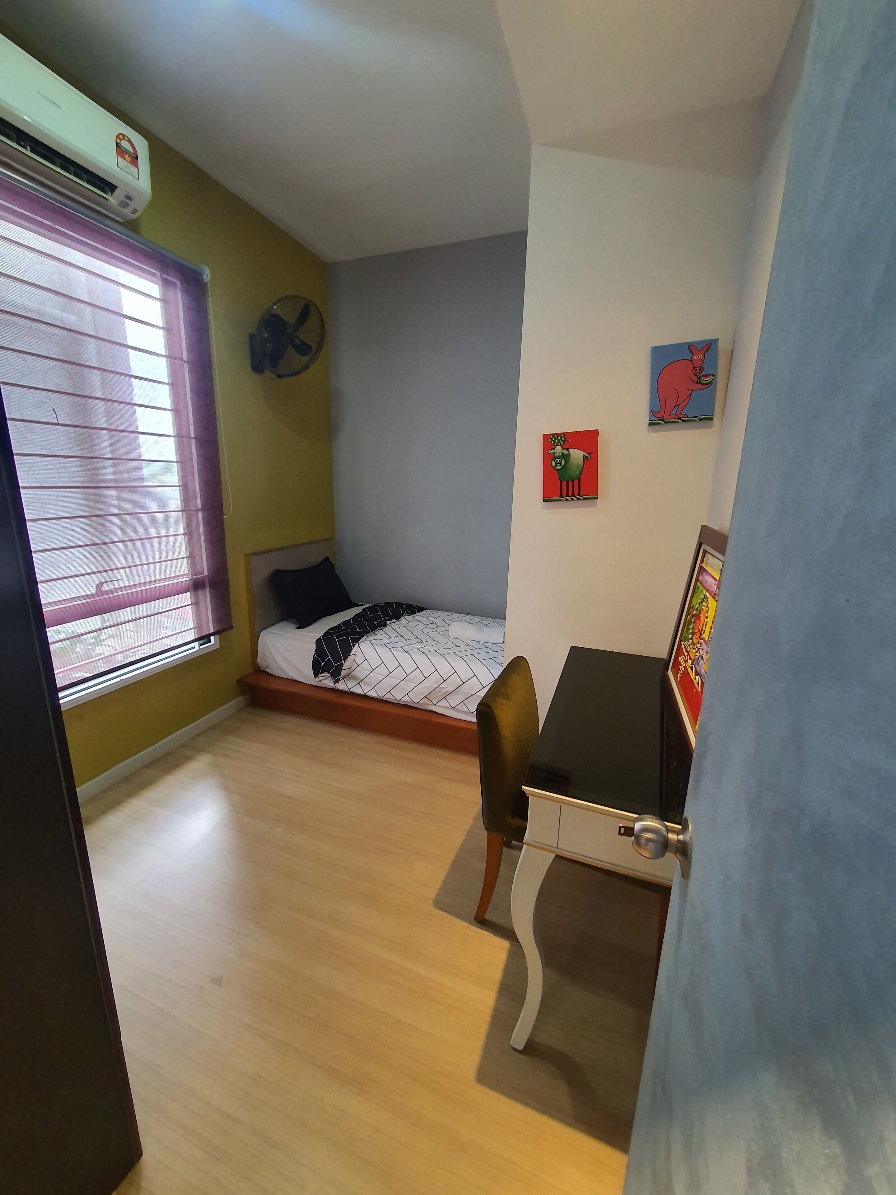 A small, well-lit room with a bed, a desk, and a chair. There is a window with blinds, a wall-mounted air conditioner, and a few wall decorations.