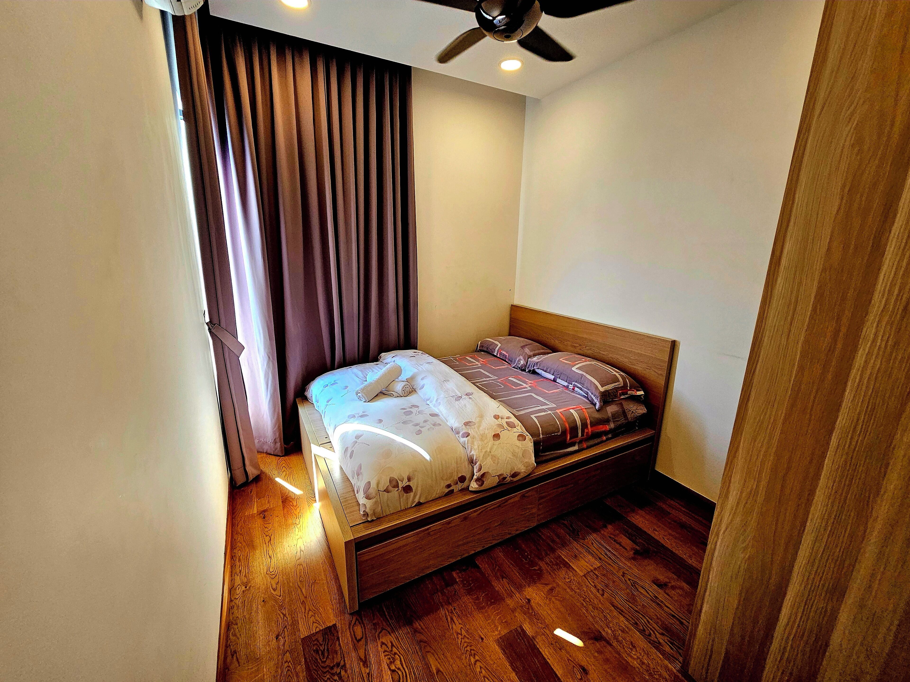 Una cama pequeña y ordenada con cabecera de madera, en una habitación con suelo de madera y ventilador de techo.