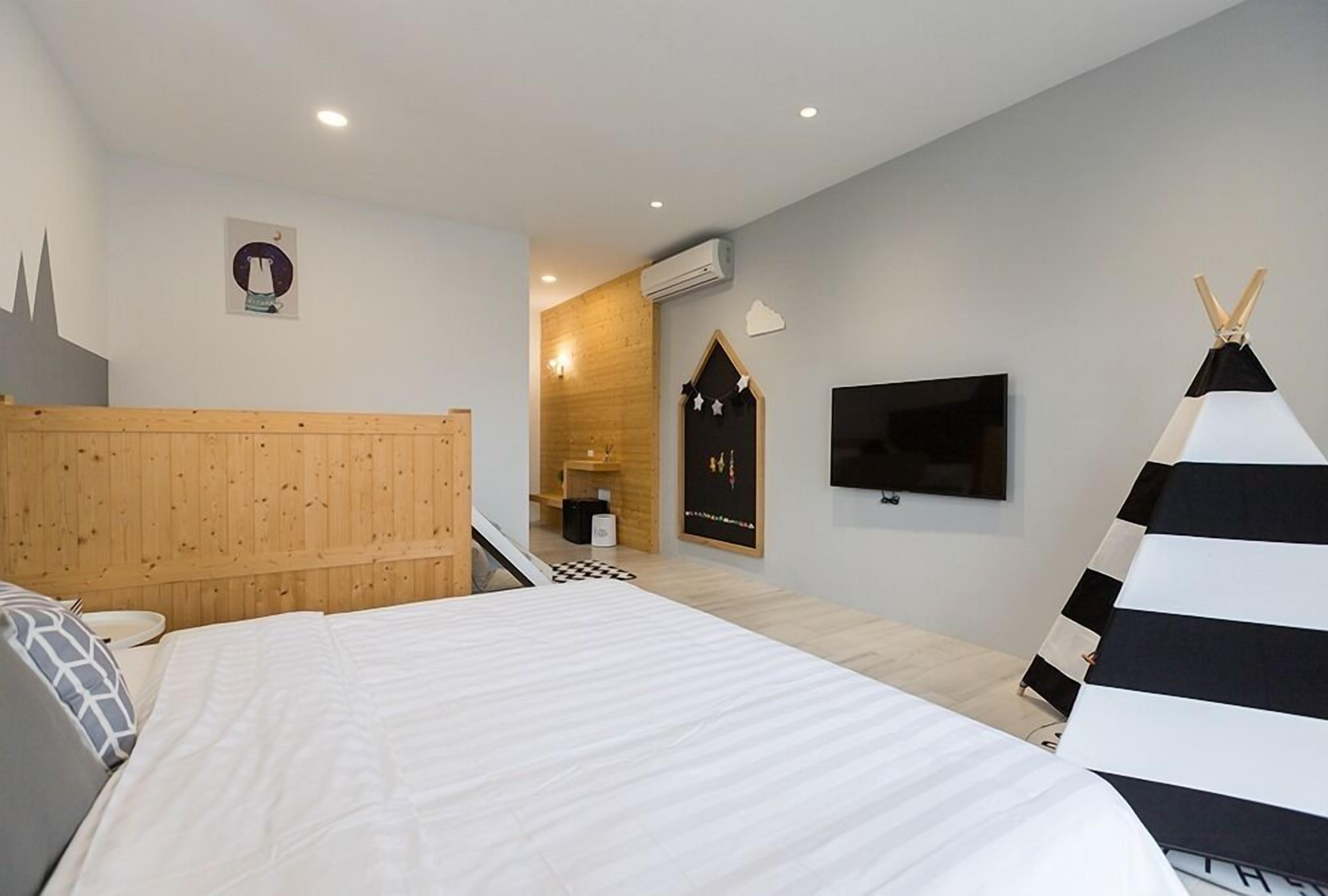 A modern bedroom with a wooden headboard, a flat-screen TV mounted on the wall, a black and white striped pillow, and a small white table with a lamp.