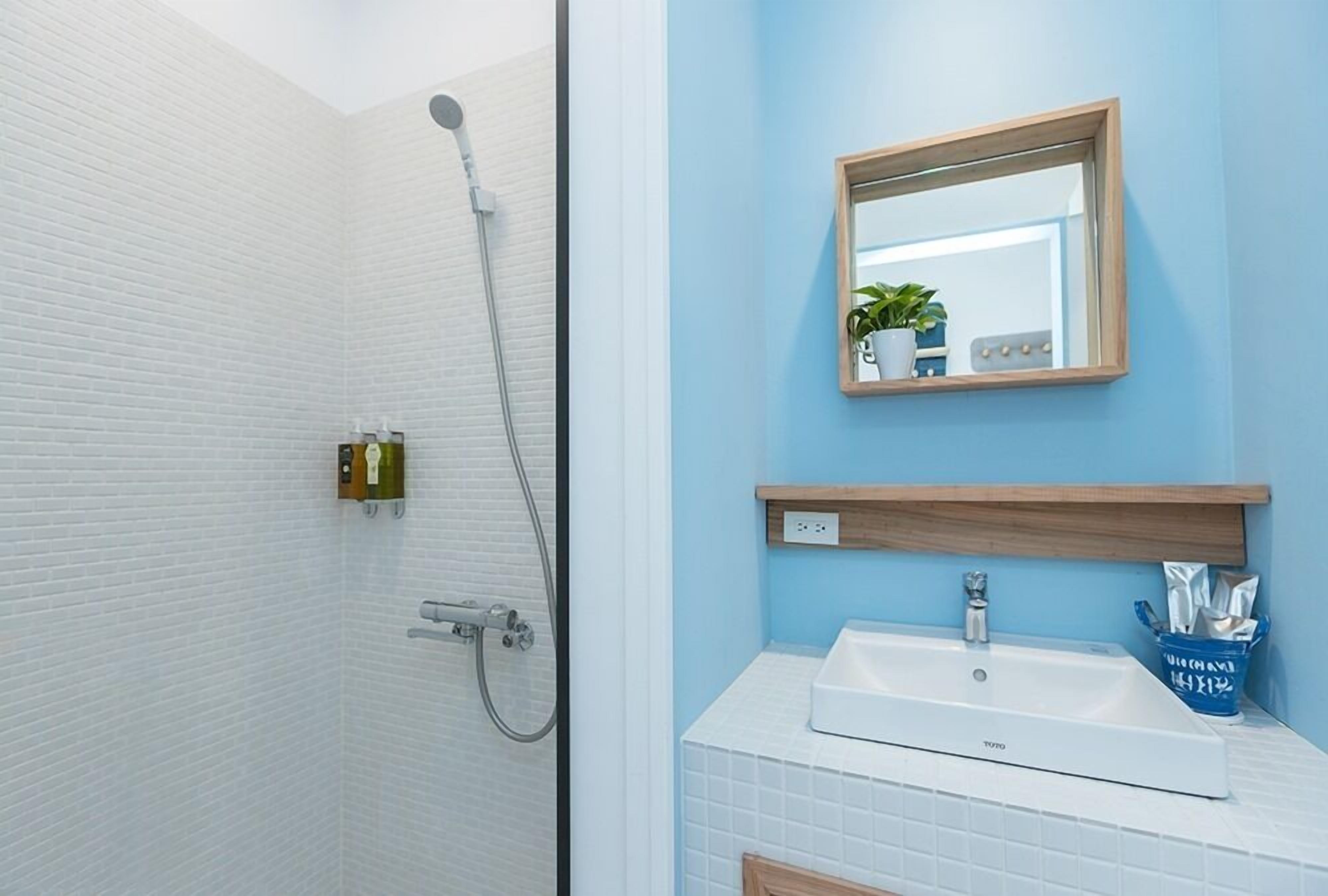 A bathroom with a white tiled shower, a white sink with a wooden shelf above, and a mirror with a plant and a blue trash bin.