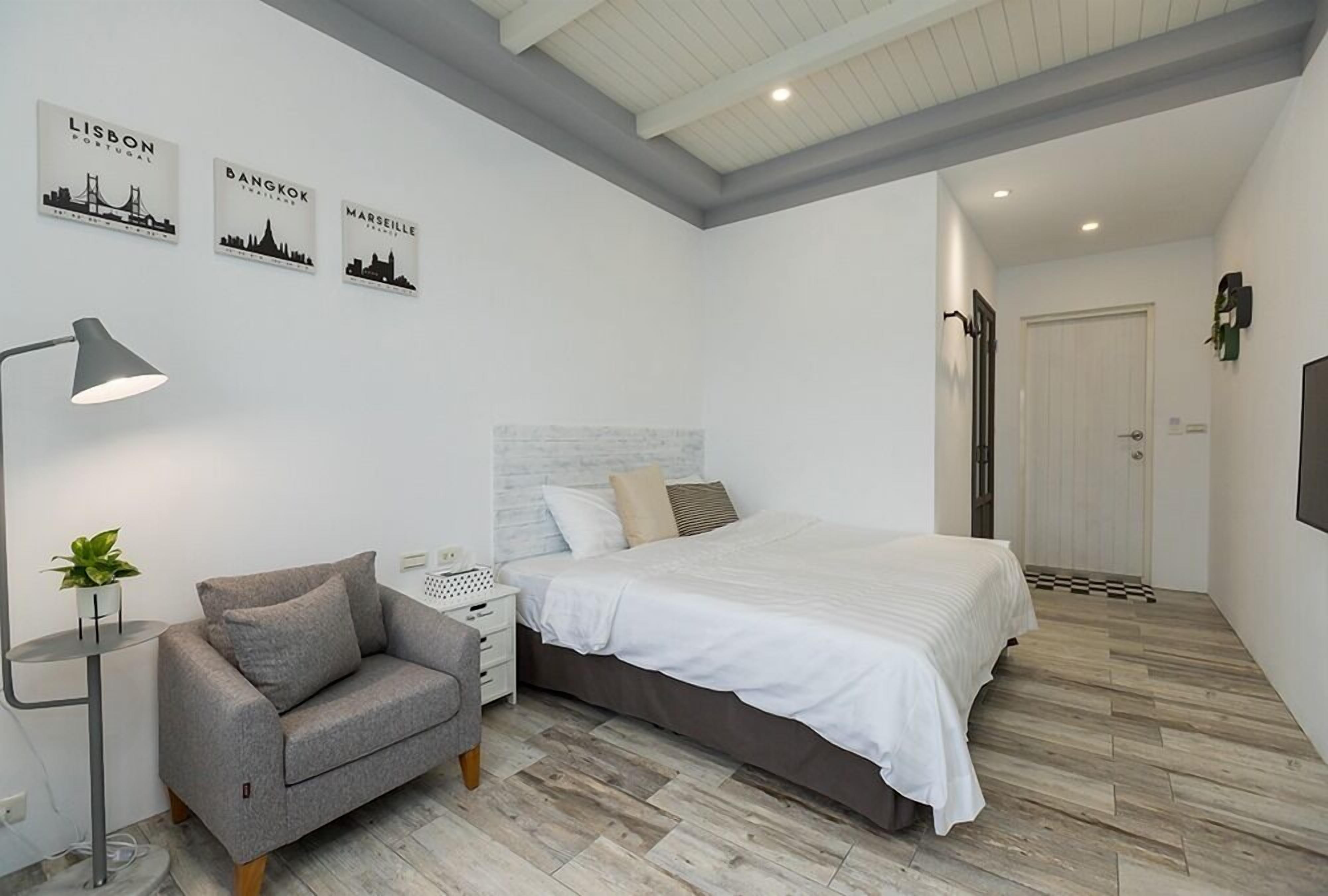 A modern bedroom with a bed, a grey sofa, a bedside table with a lamp, and wall art depicting Lisbon, Bangkok, and Marseille.