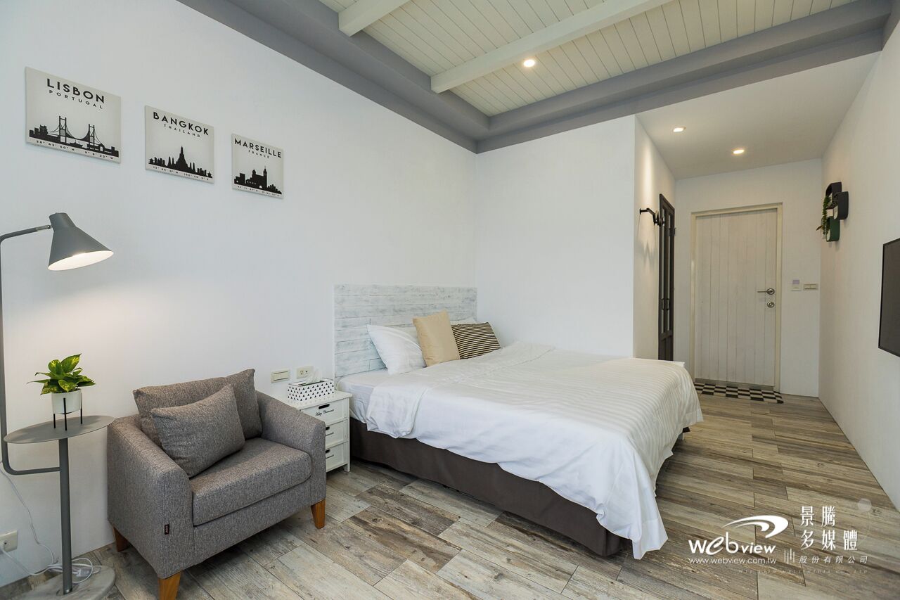 Un dormitorio moderno con una cama, un sofá gris, una mesita de noche, una lámpara y arte mural que representa Lisboa, Bangkok y Marsella.