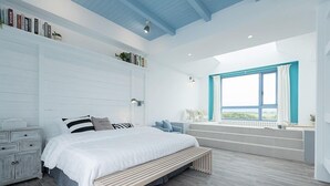 Quarto moderno com uma cama grande, cômoda branca, janela com vista e um banco embutido.