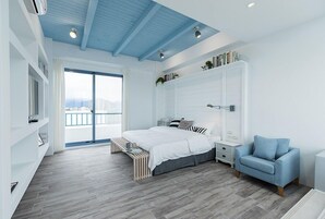 Un dormitorio moderno con una cama grande, un sillón azul, un armario blanco y vistas a la ciudad a través de un ventanal amplio.