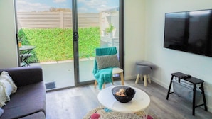 A modern living room with a sofa, a small round table with a bowl, a TV mounted on the wall, and a sliding glass door leading to an outdoor area with a green hedge and a table. - U201 Brunswick Living Top flor Balcony Close to APT n CBD FreeWifi Tram Doorstep (Brunswick West)