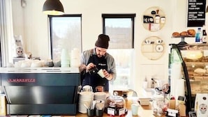 A coffee shop interior with a barista preparing a drink, a coffee machine, and various food items displayed on the counter. - U201 Brunswick Living Top flor Balcony Close to APT n CBD FreeWifi Tram Doorstep (Brunswick West)