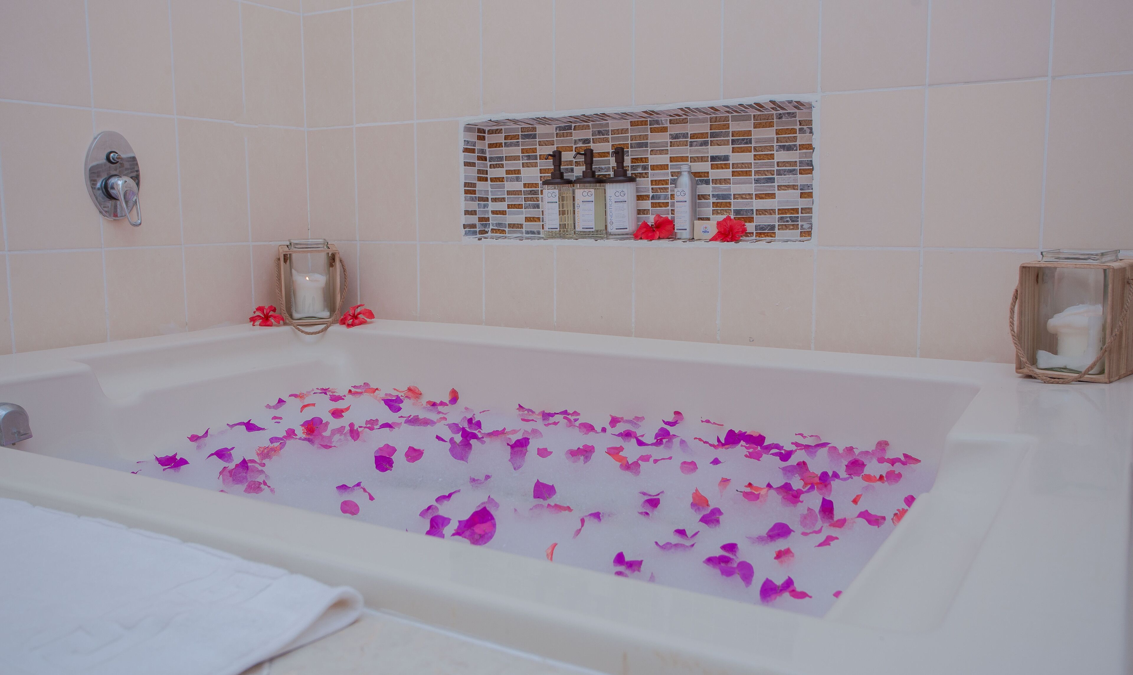 A bathtub filled with rose petals and candles, with a tiled wall and a shelf with toiletries.