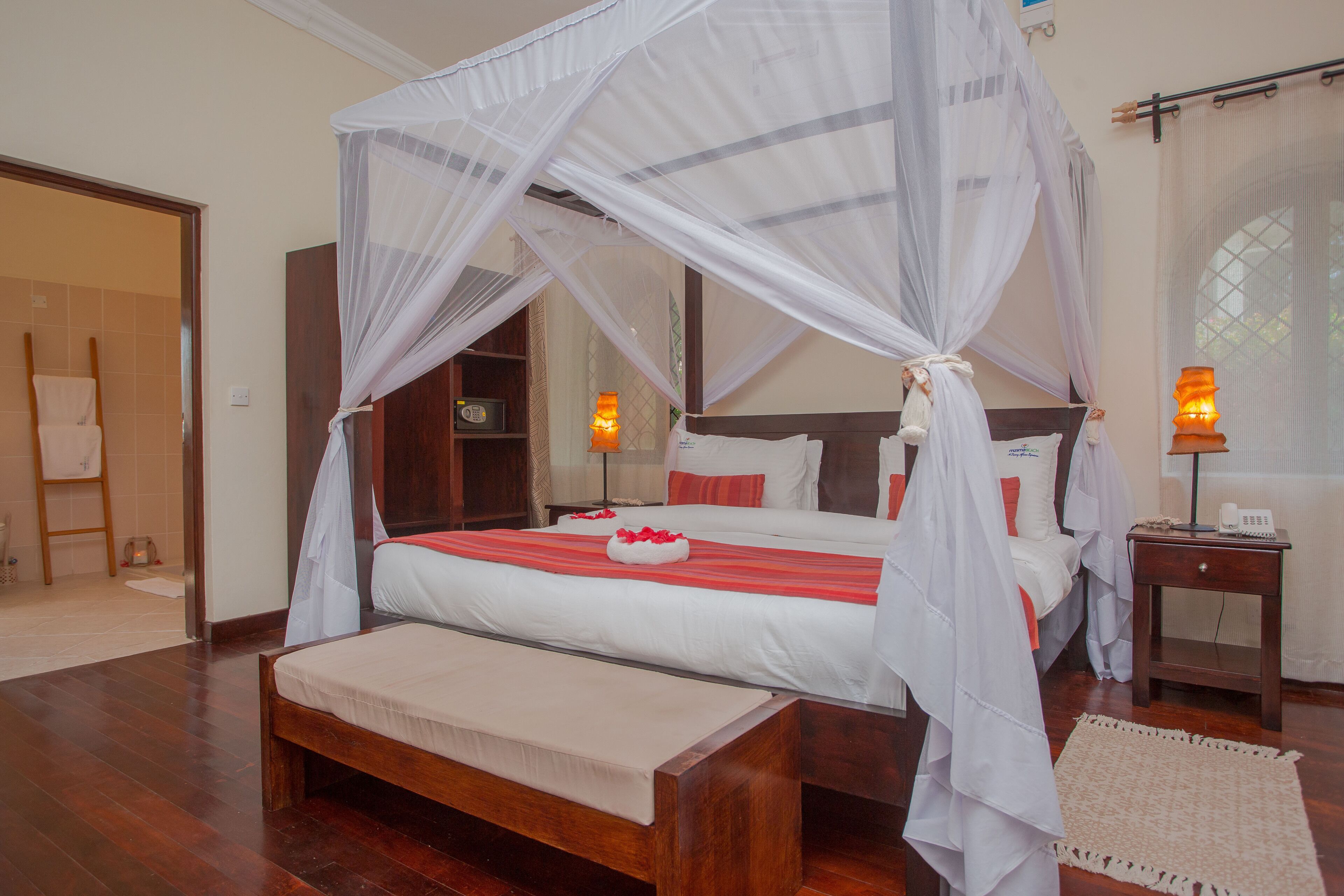 A four-poster bed with a canopy, a bedside table with a lamp, and a bench in a hotel room.