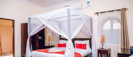 A four-poster bed with a mosquito net, red and white bedding, a bedside table with a lamp, and a window with curtains.