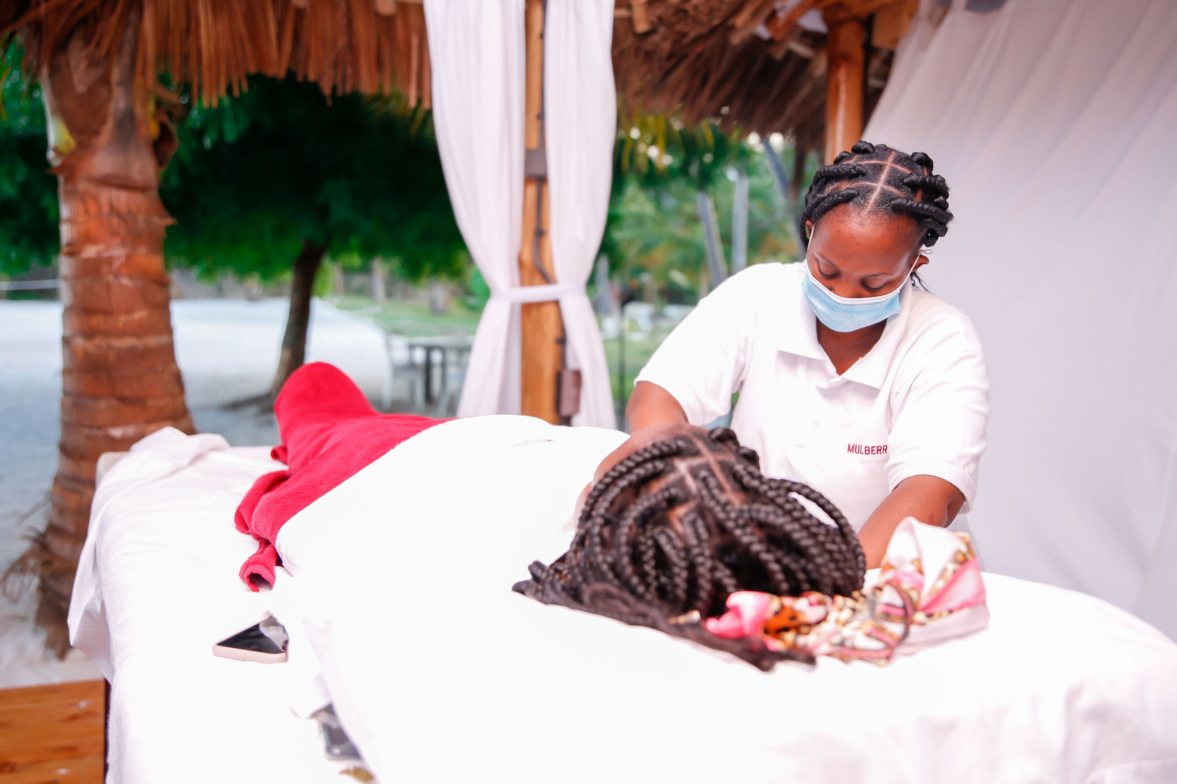 A massage therapy session in a room with a thatched roof and white drapes, a wooden floor, and a bed with a red blanket.
