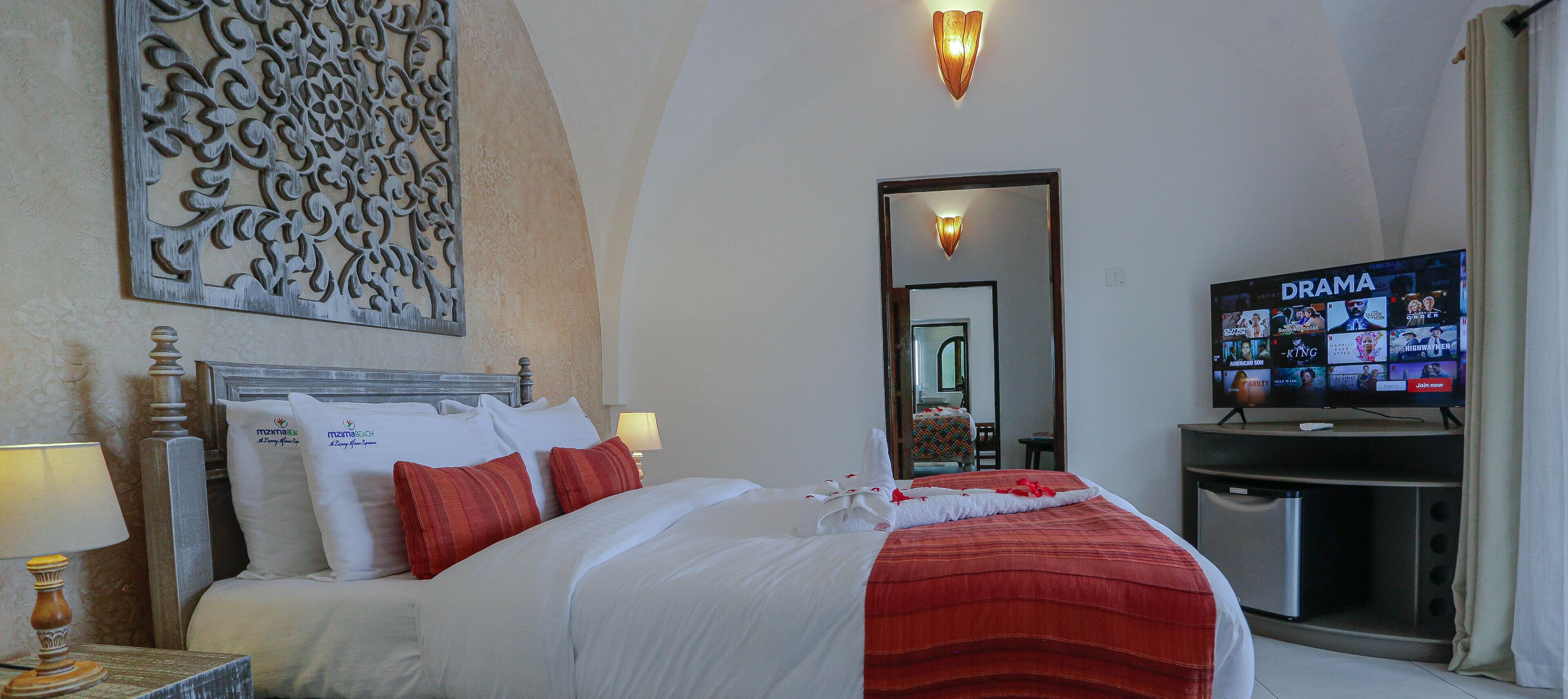 A hotel room with a bed, a TV displaying a drama channel, a wall-mounted decorative panel, and a mirror reflecting the room.