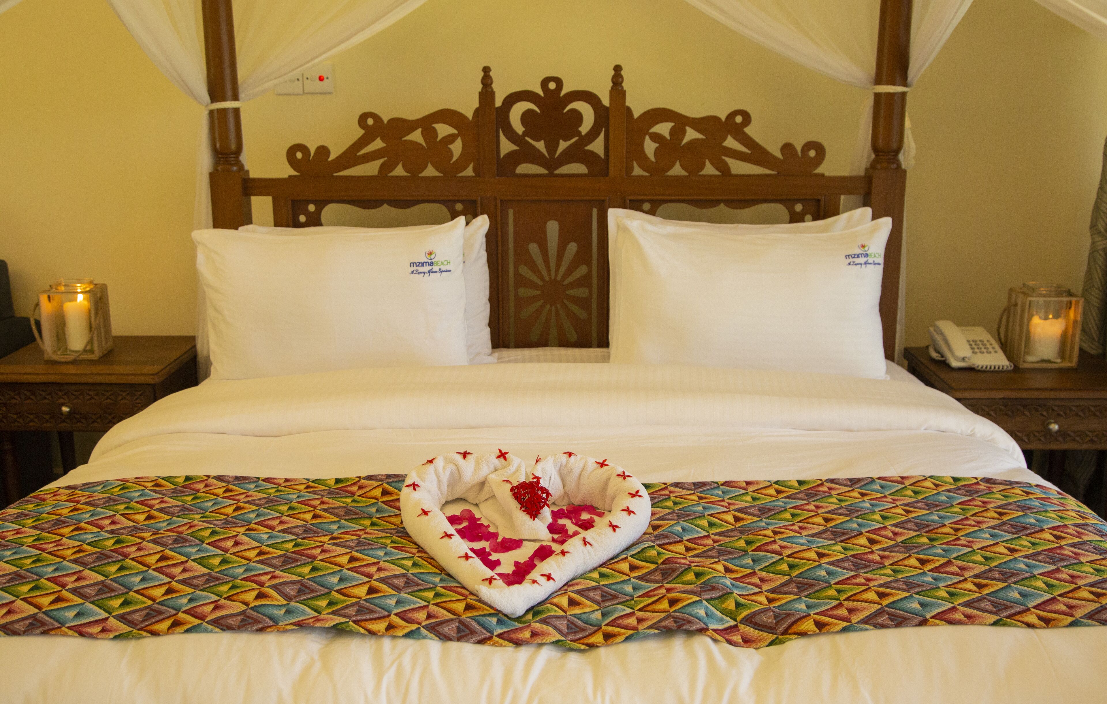 A neatly made bed with a colorful geometric-patterned bedspread, a heart-shaped decorative item, and a wooden headboard with intricate carvings.