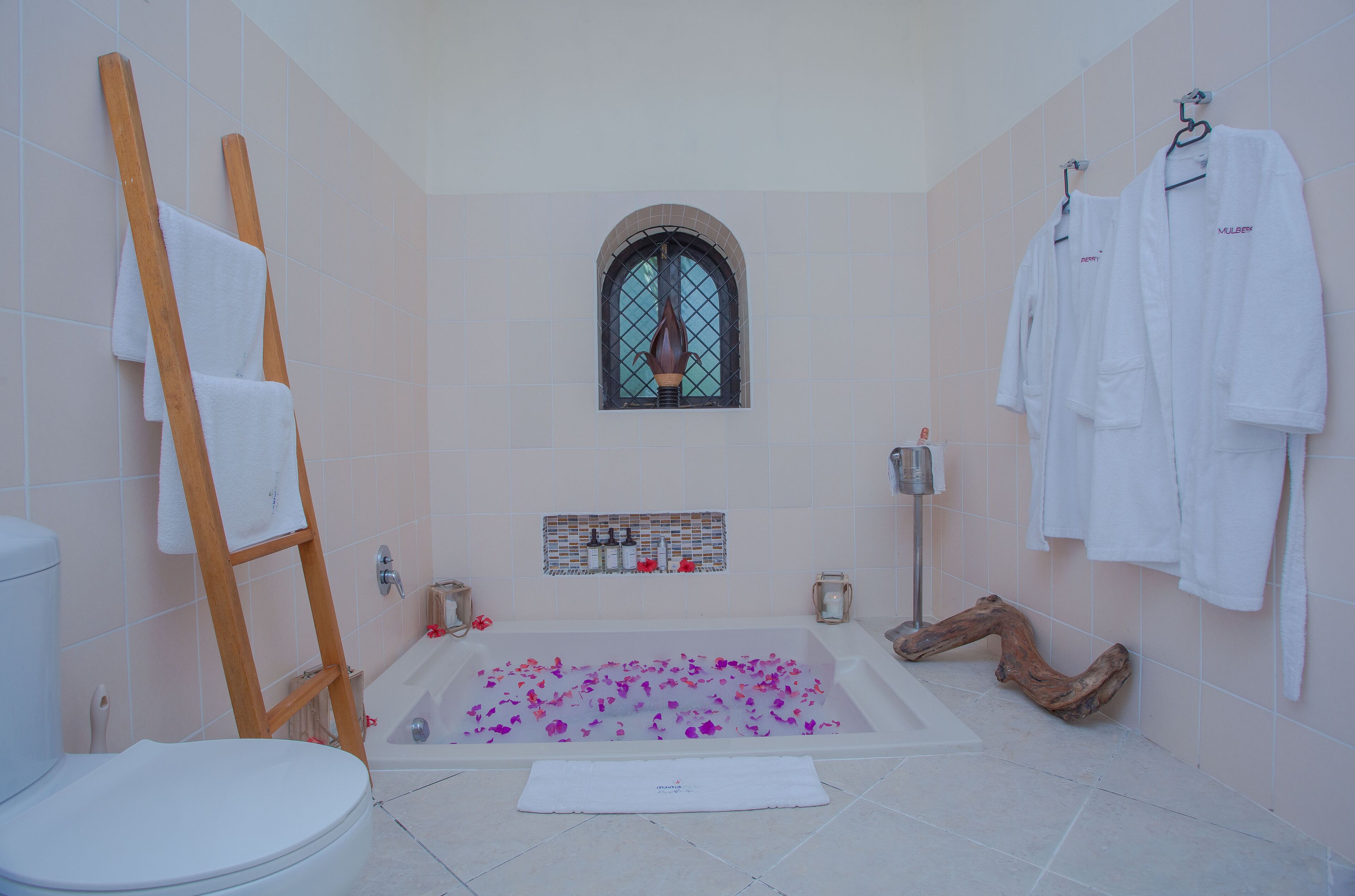 Un bagno con una vasca da bagno piena di petali di rosa, una scala di legno, una finestra e diversi asciugamani bianchi appesi a ganci.