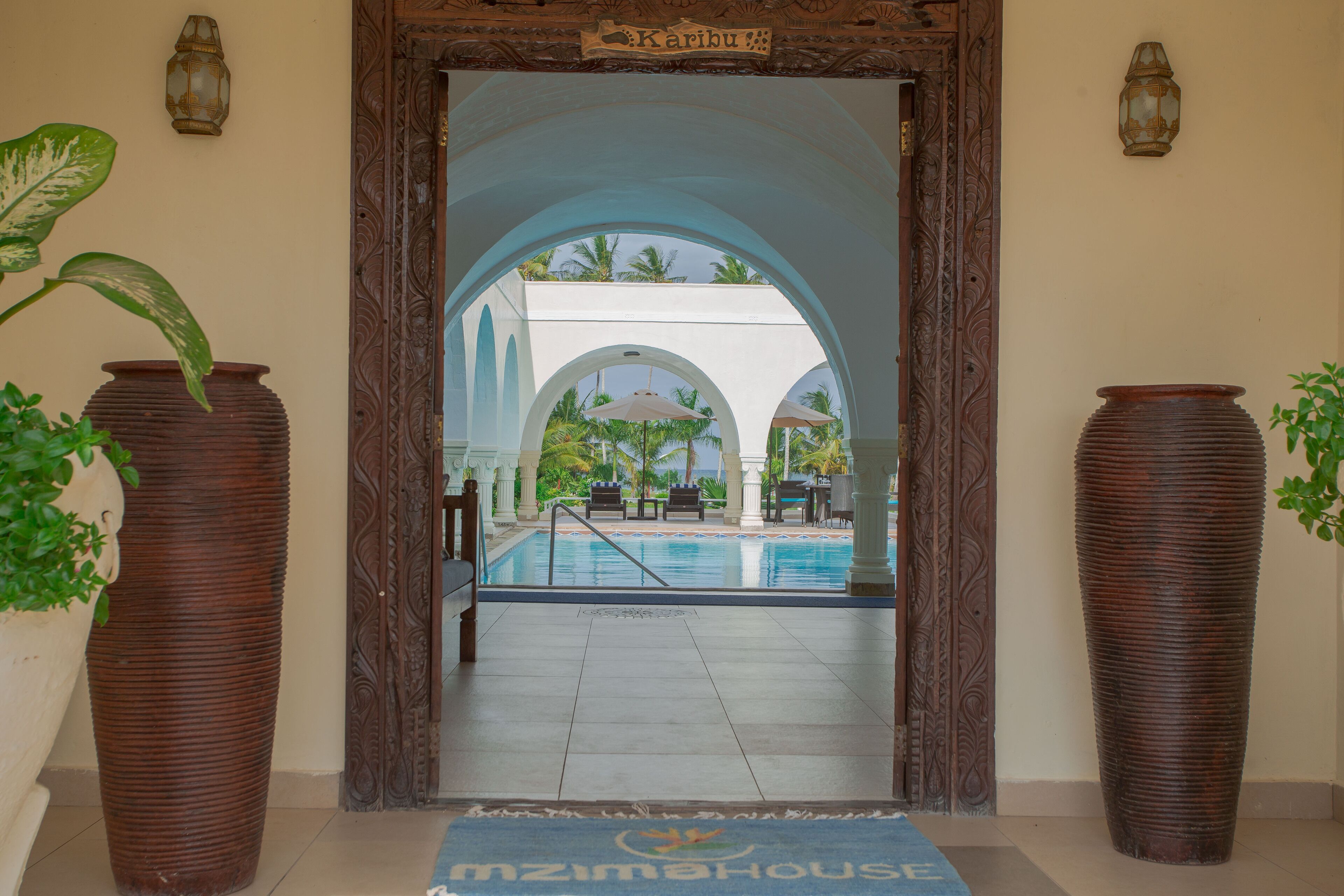 A hotel entrance with a pool area visible through an arched doorway, featuring large potted plants and a sign above the entrance.