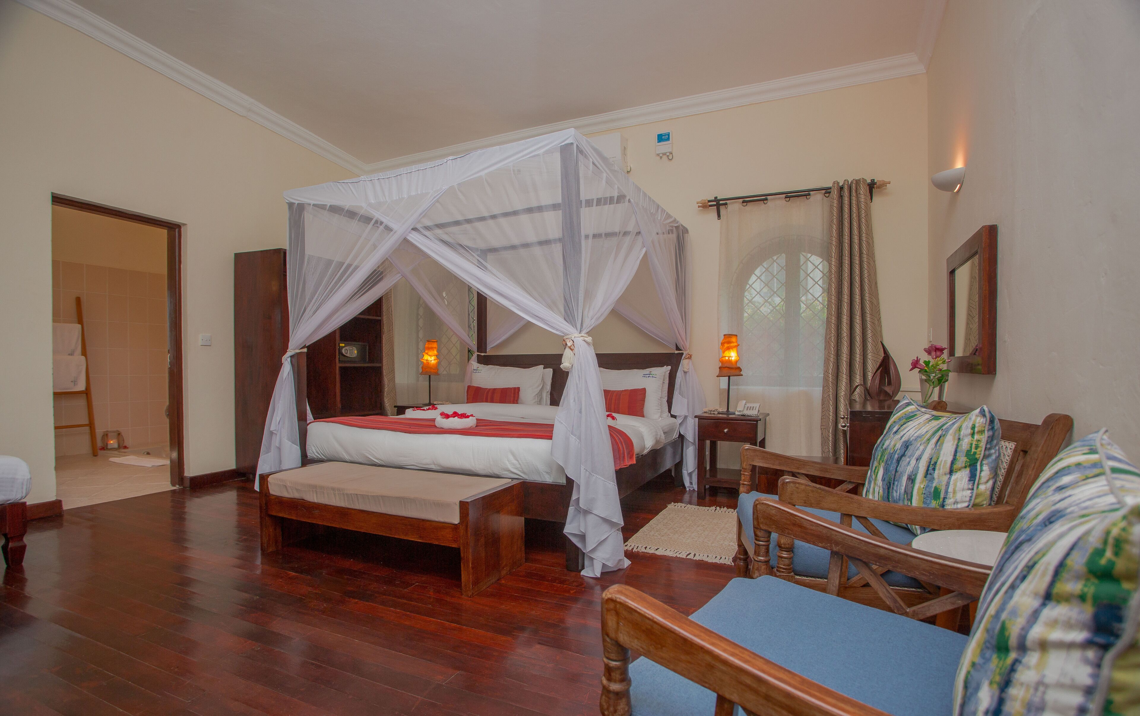 A hotel room with a four-poster bed, a sitting area with a chair and a small table, and a large window with curtains.