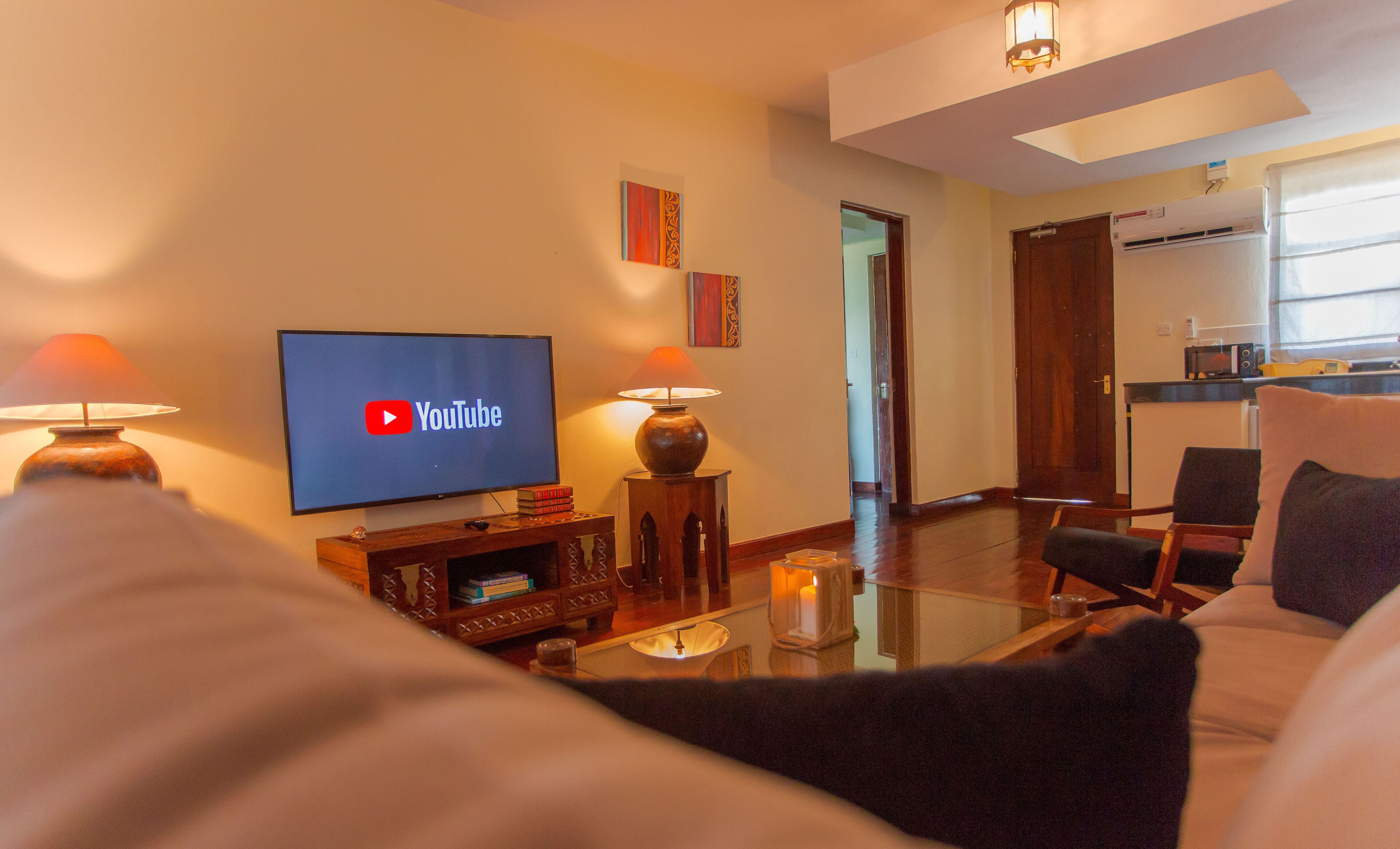 A living room with a flat-screen TV displaying the YouTube logo, a coffee table, a sofa, and a dining area with chairs.