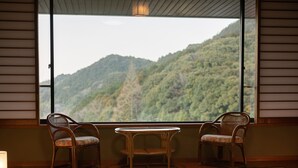 A room with tatami flooring, a low table, two chairs, and a view of a forested mountain. - Yamamura Annex (Nagato)