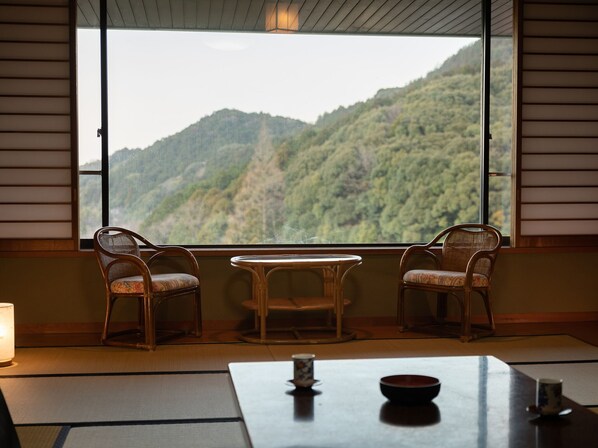 Une pièce avec un sol recouvert de tatamis, une table basse, deux chaises et une vue sur une montagne boisée.