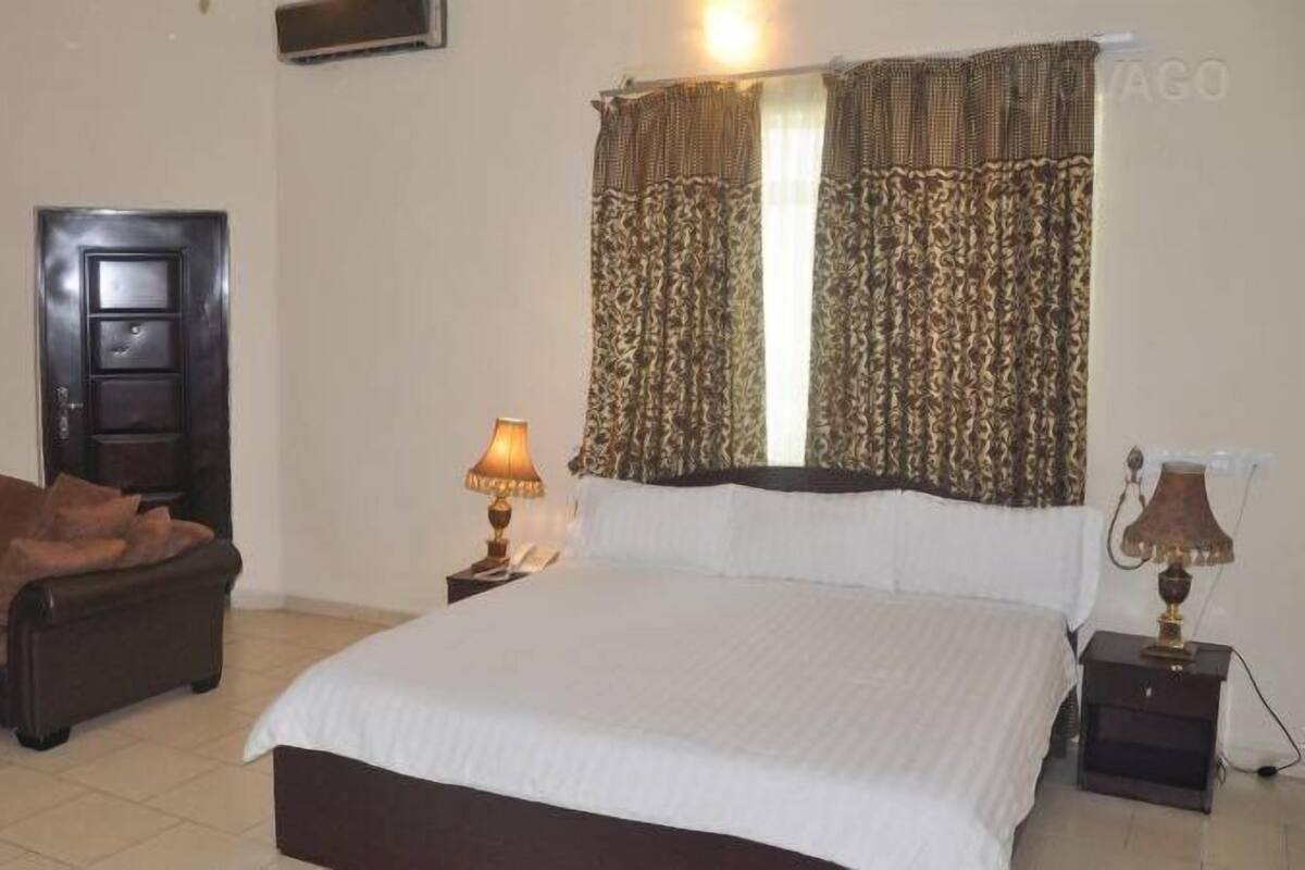 A hotel room with a bed, a chair, a nightstand with a lamp, and a door.