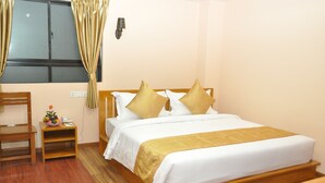 Habitación de hotel ordenada con una cama de madera, una mesita de noche con lámpara, una pequeña mesa con frutero y una silla.