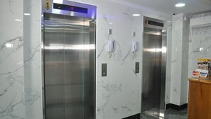 Dos ascensores con paredes de mármol y pantalla digital, un teléfono público y un mostrador de madera con logotipos de Visa y Mastercard.