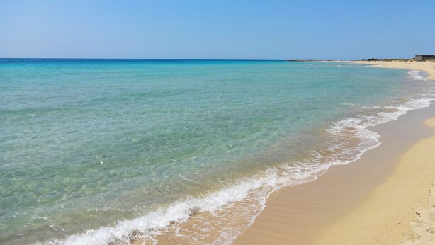 Een strand met helder turquoise water, een zandstrand en een gebouw in de verte.