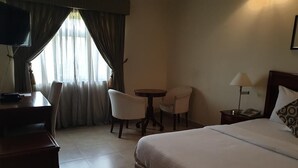 A hotel room with a bed, a desk, two chairs, a small table, a lamp, and a TV. - Newton Hotel Festac (Lagos)