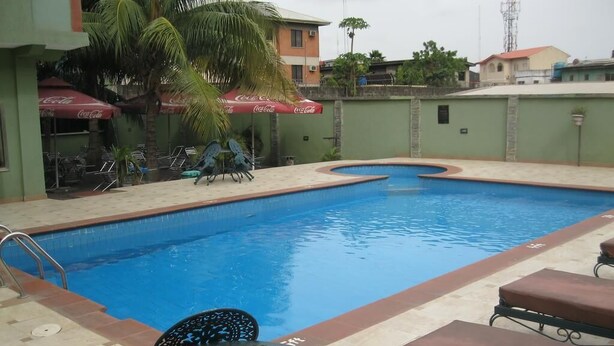 A swimming pool with lounge chairs and tables, surrounded by a low wall and palm trees. - Newton Hotel Festac (Lagos)