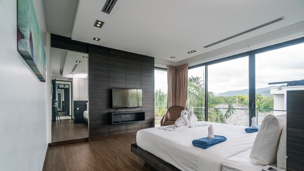 Ein modernes Hotelzimmer mit einem großen Bett, einem Flachbildfernseher, einer Sitzecke mit einem Sessel und einem Balkon mit Blick auf Grünflächen.