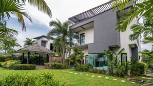 Ein modernes, zweistöckiges Haus mit einer überdachten Terrasse, umgeben von üppiger Vegetation und Palmen.