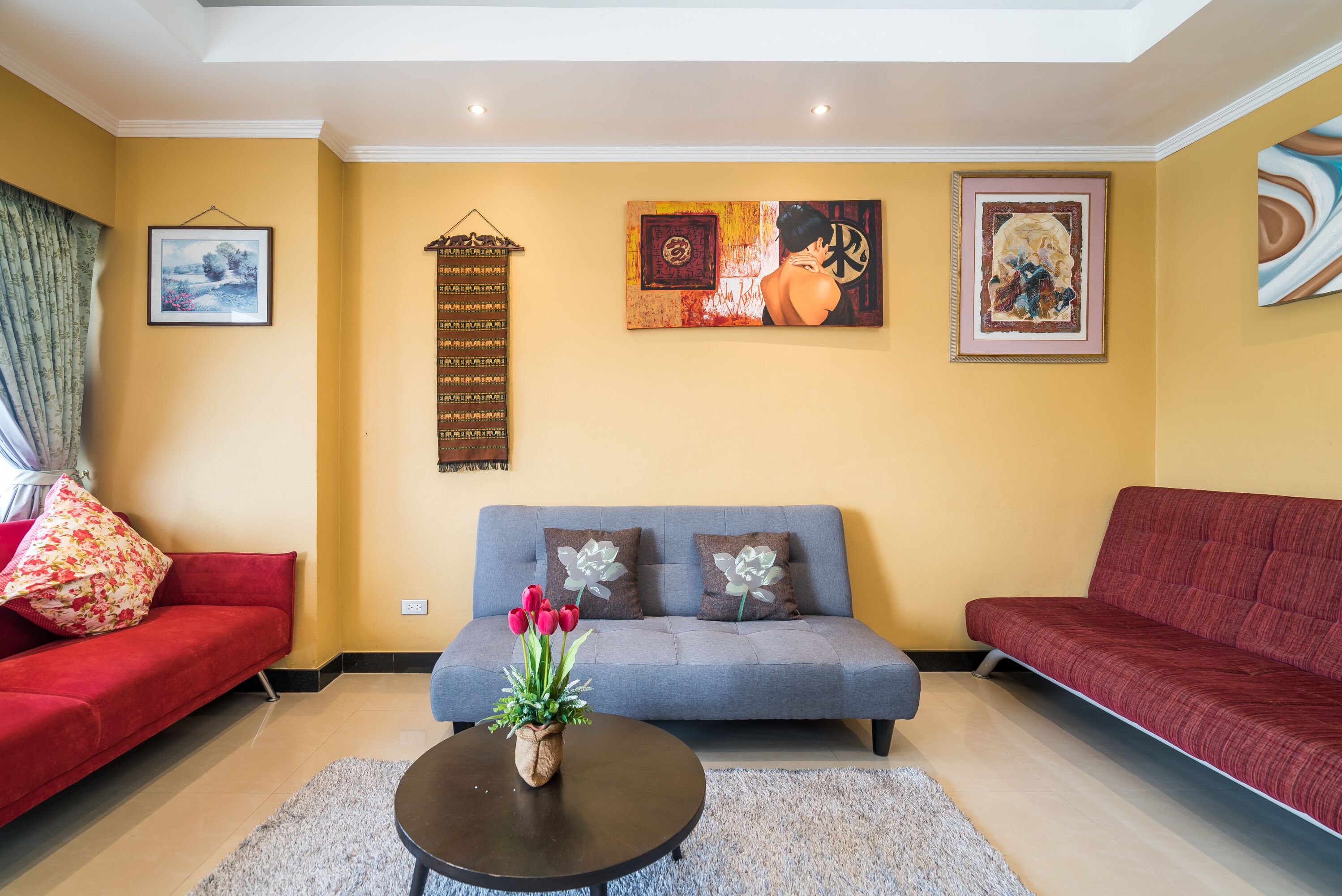Uma sala de estar com um sofá cinzento, um sofá vermelho e uma mesa de centro com um arranjo de flores. Há três quadros na parede e uma janela com cortinas.