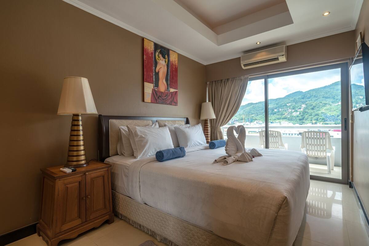 A hotel room with a large bed, a painting on the wall, and a view of the mountains through the window.
