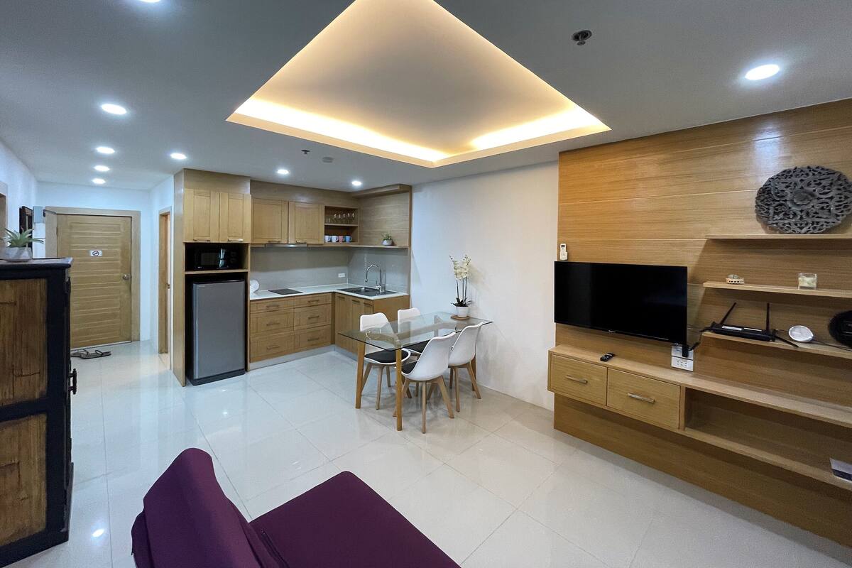 A modern kitchen with a dining area, a TV, and a flat-screen TV on a wooden wall.