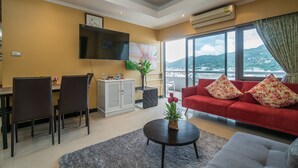 A modern living room with a red sofa, a grey armchair, a dining table with chairs, a flat-screen TV, and a large window with a view of a cityscape and mountains. - Patong Tower by Lofty (Patong)