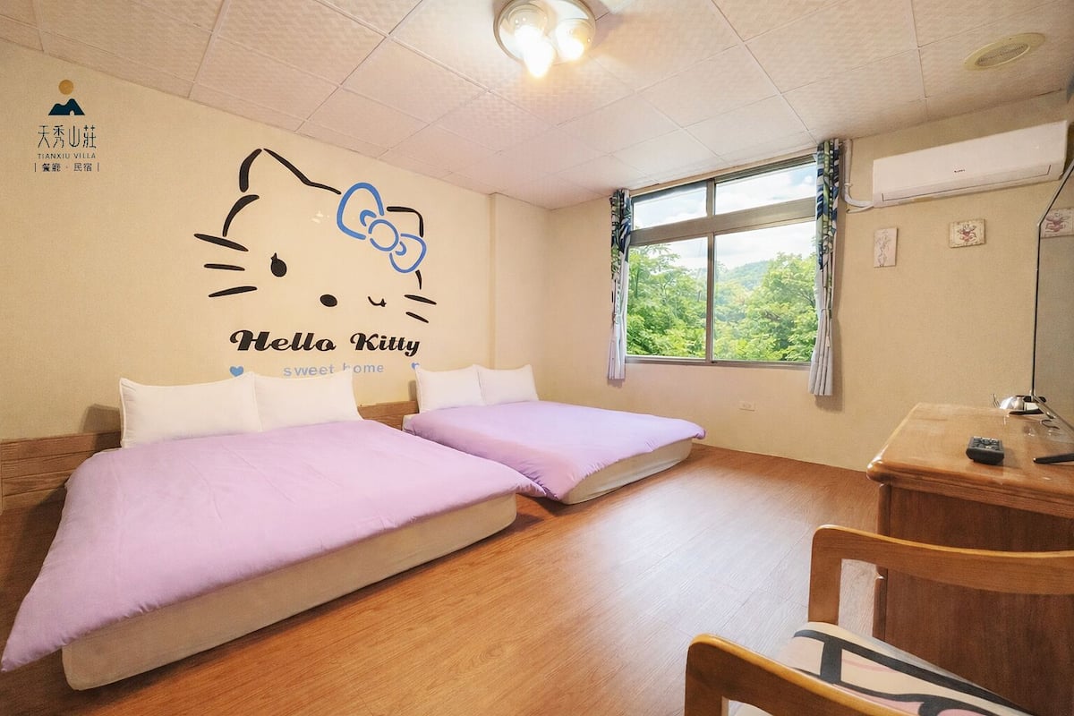 A hotel room with two beds, a desk, a chair, a window with a view of greenery, and a Hello Kitty wall decal.