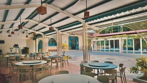 Un comedor interior con mesas y sillas redondas, una zona de estar exterior cubierta con vista a la calle, y un pasillo techado con techo de malla.